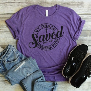 May include: Purple t-shirt with a black circle graphic that says "Saved by Grace through Faith" with a cross and the scripture reference "Eph. 2:8".
