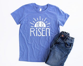 Puede incluir: Una camiseta azul de manga corta con un gráfico de sol blanco y el texto "He is Risen".