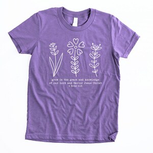 May include: Purple short-sleeved t-shirt with a white floral design and the text "grow in the grace and knowledge of our Lord and Savior Jesus Christ 2 Peter 3:10".