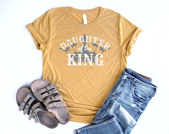 May include: A mustard yellow t-shirt with the text "Daughter of the King" and a gray crown graphic. The shirt is paired with blue jeans and brown sandals.
