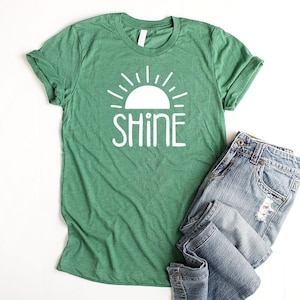 May include: Green t-shirt with a white sun and the word "shine" printed on it. The t-shirt is folded and laid on a white surface next to a pair of blue jeans.
