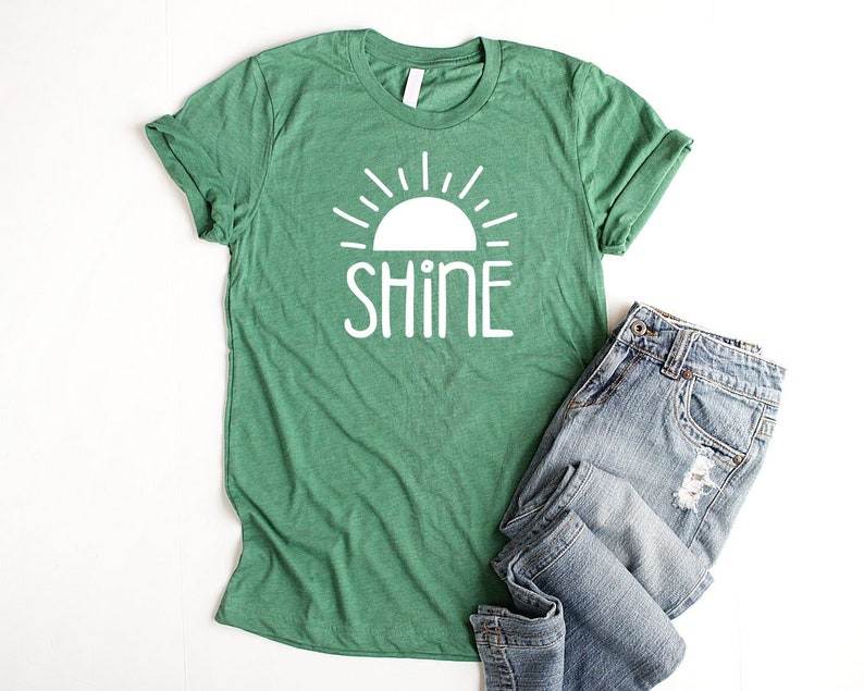 May include: Green t-shirt with a white sun and the word "shine" printed on it. The t-shirt is folded and laid on a white surface next to a pair of blue jeans.