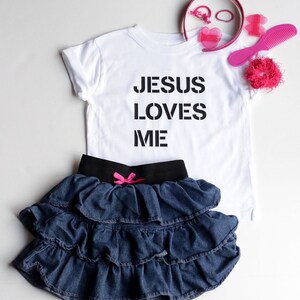 May include: White t-shirt with black text that reads "JESUS LOVES ME". A denim skirt with ruffles and a pink bow is also pictured.