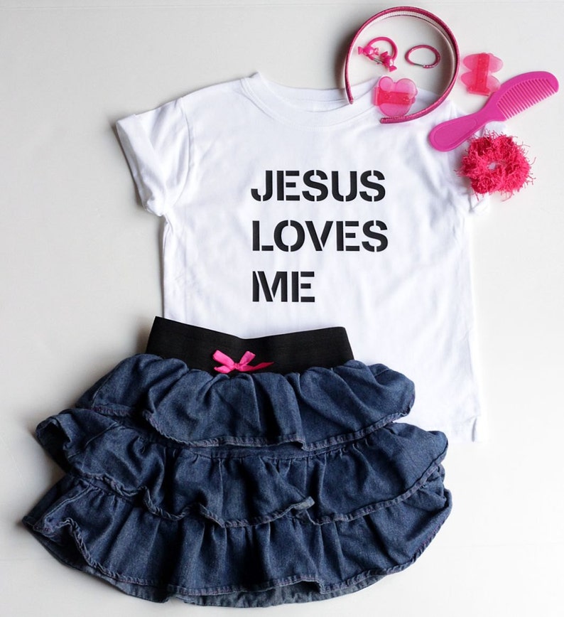 May include: White t-shirt with black text that reads "JESUS LOVES ME". A denim skirt with ruffles and a pink bow is also pictured.