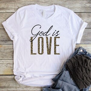 May include: White t-shirt with the text "God is Love" printed in black. The word "Love" is printed in a leopard print pattern.