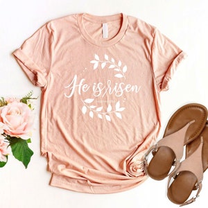 Puede incluir: Una camiseta de color melocot&oacute;n con el texto "He is risen" en letras blancas con una corona de flores alrededor del texto. La camiseta est&aacute; sobre una superficie blanca con un par de sandalias marrones.
