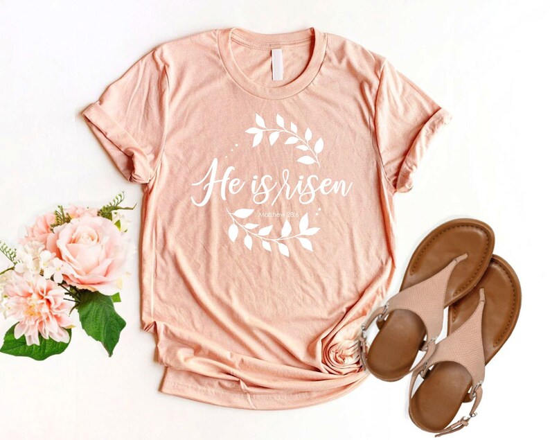 Puede incluir: Una camiseta de color melocot&oacute;n con el texto "He is risen" en letras blancas con una corona de flores alrededor del texto. La camiseta est&aacute; sobre una superficie blanca con un par de sandalias marrones.