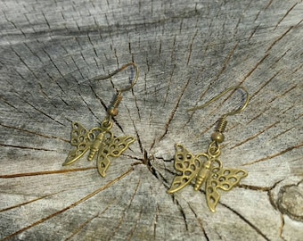 May include: A pair of gold-toned butterfly earrings with intricate designs. The earrings are hanging from a wooden surface.