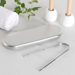 May include: Two silver tongue scrapers in a silver metal case. The scrapers are made of stainless steel and have a long, thin handle.
