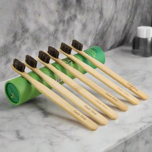 May include: Six bamboo toothbrushes with black bristles and the text "Gaia Guy" printed on the handles. The toothbrushes are arranged in a row on a white marble surface.