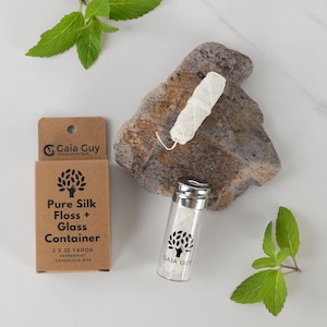 May include: A glass container of white floss with a silver lid, next to a cardboard box labeled "Pure Silk Floss + Glass Container." The box and container have a tree logo and the text "Gaia Guy." A piece of floss sits on a rock, with mint leaves scattered around.