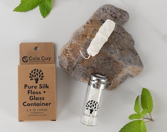 May include: A glass container of white floss with a silver lid, beside a cardboard box labelled "Pure Silk Floss + Glass Container." The box and container have a tree logo and the text "Gaia Guy." A piece of floss sits on a rock, with mint leaves scattered around.