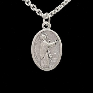 May include: Silver oval pendant necklace with a detailed relief of a robed figure with outstretched arms, labelled "S. GENEVIEVE." The pendant hangs from a delicate silver chain. The background is black.