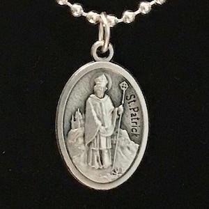 May include: Silver oval pendant necklace with a depiction of Saint Patrick holding a staff. The pendant has the text "St. Patrick" inscribed on the side.