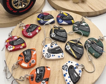 May include: A collection of miniature racing helmet earrings in various colors including red, orange, white, black, blue, and green. Each helmet features a black visor and a unique racing number. The earrings are displayed on a circular wooden base.