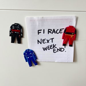 May include: A white note with black handwritten text that reads "F1 Race Next Weekend."  Three small toy race car driver figurines are attached to the note with magnets. One figurine is black and red, one is red and yellow, and one is blue and white.