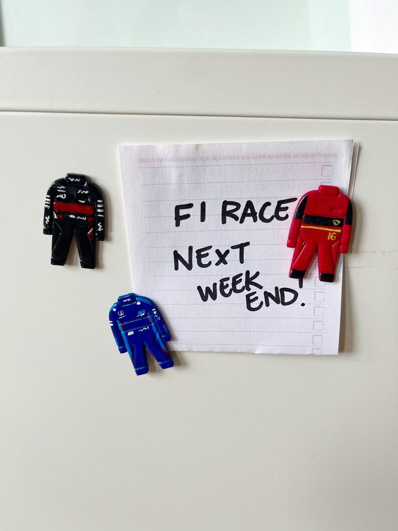 May include: A white note with black handwritten text that reads "F1 Race Next Weekend."  Three small toy race car driver figurines are attached to the note with magnets. One figurine is black and red, one is red and yellow, and one is blue and white.