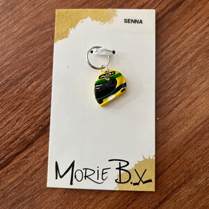 May include: A yellow, green, and black racing helmet charm with a silver key ring. The charm is on a white card with the text "SENNA" and "MORIE B.X."
