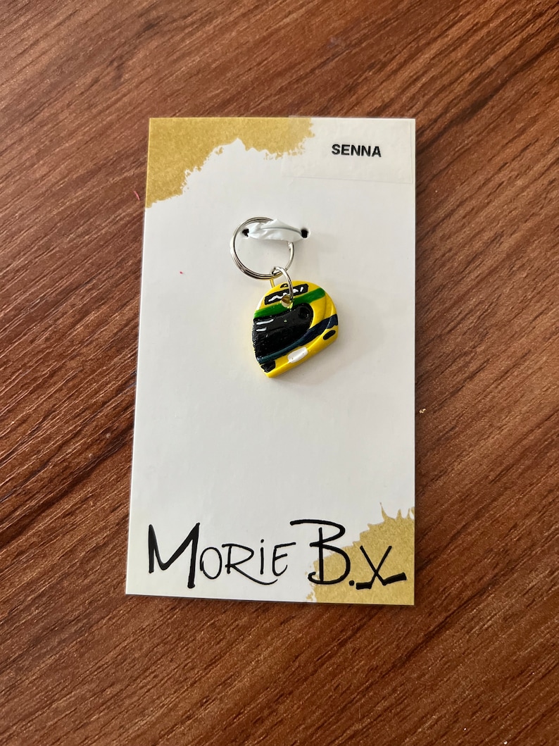 May include: A yellow, green, and black racing helmet charm with a silver key ring. The charm is on a white card with the text "SENNA" and "MORIE B.X."