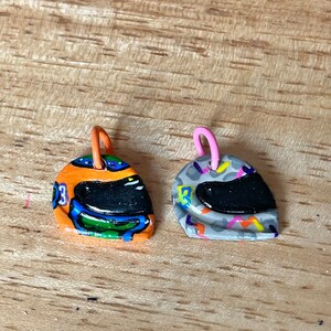 May include: Two small, colorful, hand-painted, ceramic helmet charms. One is orange, blue, green, and black. The other is gray, white, orange, and black.