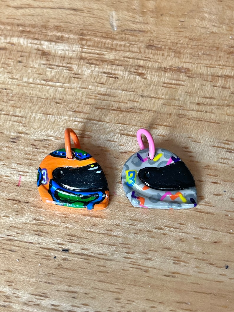May include: Two small, colorful, hand-painted, ceramic helmet charms. One is orange, blue, green, and black. The other is gray, white, orange, and black.