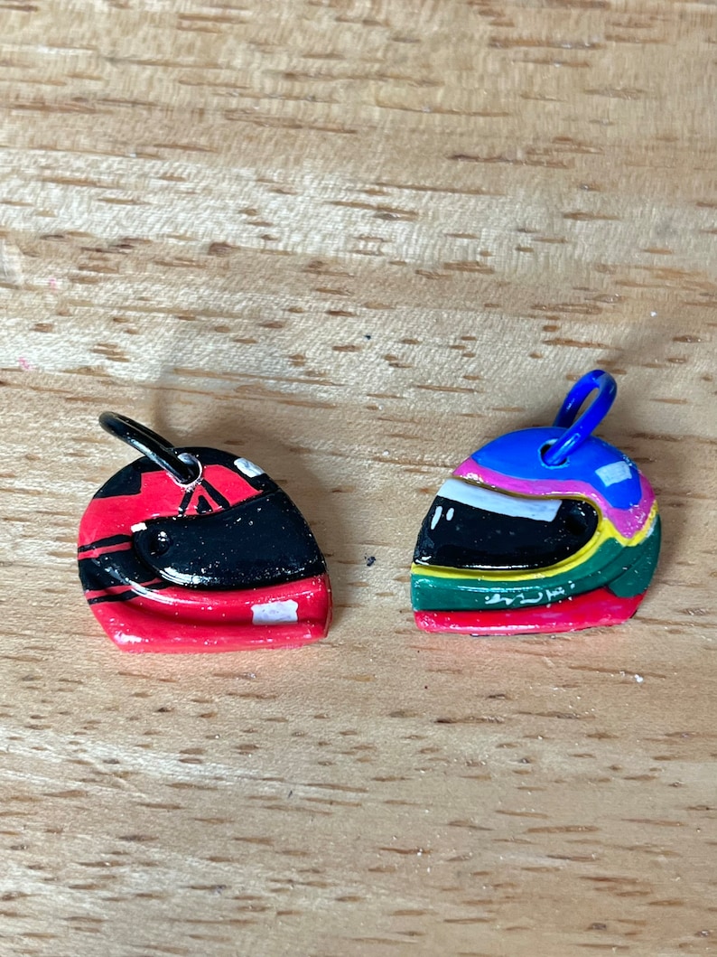 May include: Two small, hand-painted racing helmet charms. One is red with black and white stripes, the other is green, blue, and yellow with a white stripe.