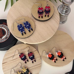Può includere: Orecchini a tema hockey in vari colori delle squadre, tra cui rosso, blu, giallo e arancione. Ogni orecchino presenta un design di giocatore di hockey in miniatura con ganci color argento. Gli orecchini sono esposti su piccoli cerchi di legno.