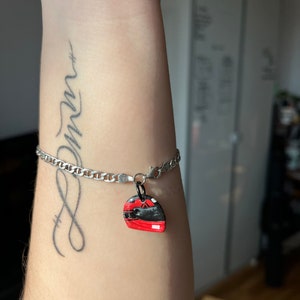 May include: A silver chain bracelet with a red and black helmet charm. The bracelet is on a person's wrist, and the charm is hanging from the chain. The person has a tattoo on their wrist that says "dream".