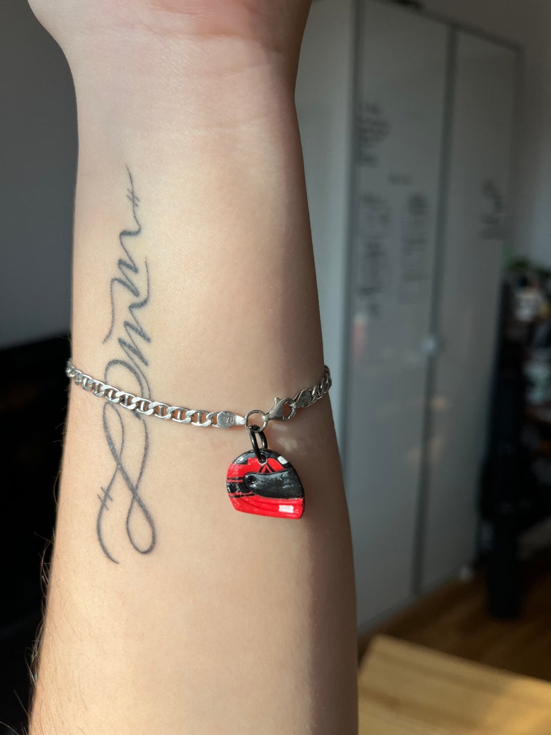 May include: A silver chain bracelet with a red and black helmet charm. The bracelet is on a person's wrist, and the charm is hanging from the chain. The person has a tattoo on their wrist that says "dream".