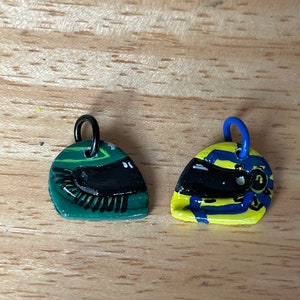 May include: Two small, painted, ceramic charms shaped like racing helmets. One is green with black details and the other is yellow with blue and black details.