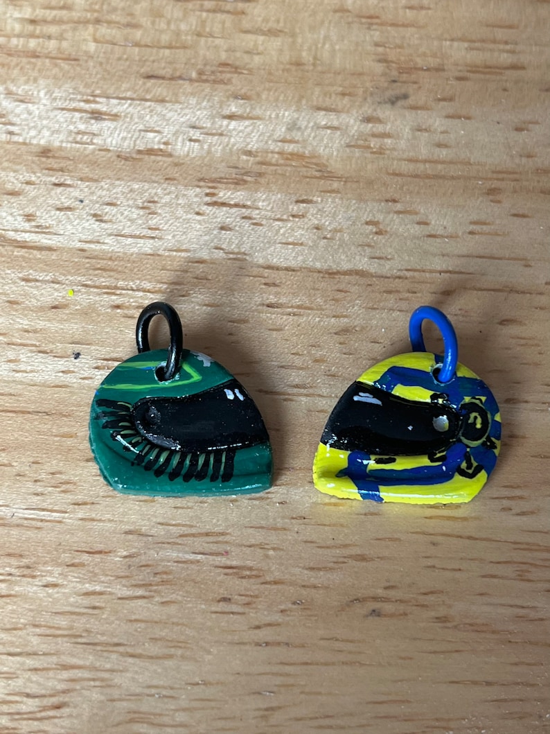 May include: Two small, painted, ceramic charms shaped like racing helmets. One is green with black details and the other is yellow with blue and black details.