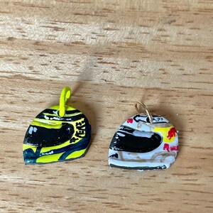 May include: Two small, painted metal helmet charms. One is yellow with black and white details, the other is white with black, red, and yellow details.