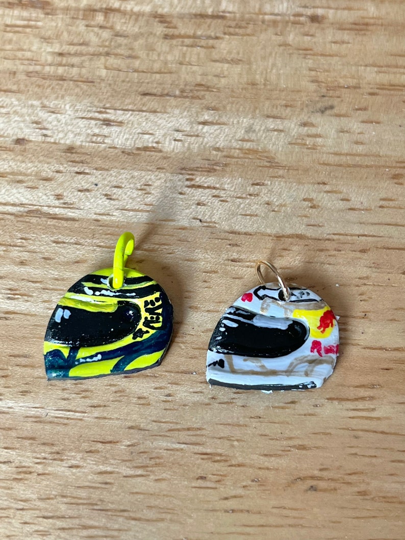 May include: Two small, painted metal helmet charms. One is yellow with black and white details, the other is white with black, red, and yellow details.