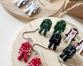May include: A collection of dangle earrings shaped like miniature racing driver suits in various colors including red, black, white, green, and orange. Each earring features a metal hook and a detailed figurine of a racing jumpsuit with painted accents.