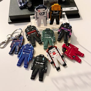 May include: A collection of nine miniature racing driver suits in various colors, including red, blue, green, black, white, and yellow. Each suit has a unique design and features a number on the chest.
