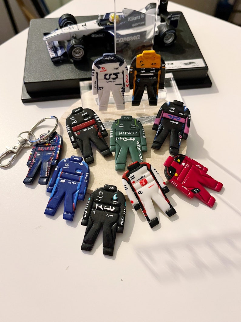 May include: A collection of nine miniature racing driver suits in various colors, including red, blue, green, black, white, and yellow. Each suit has a unique design and features a number on the chest.