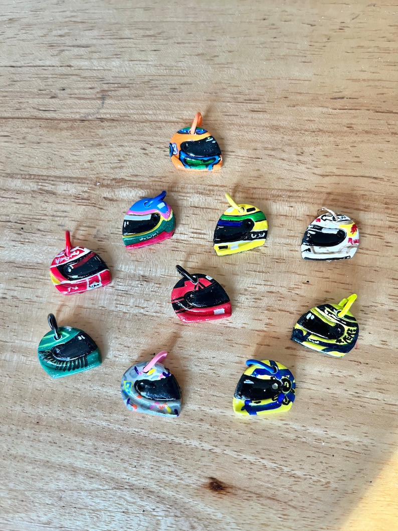 May include: A collection of ten miniature racing helmets, each with a unique design and color scheme. The helmets are made of plastic and have a small loop at the top for hanging.