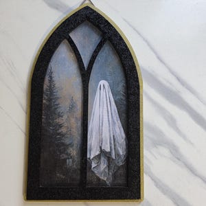 May include: A gothic-style wall hanging featuring a painting of a ghost in a window. The black frame has a gold border. The painting depicts a white ghost in front of a forest scene with a starry sky.