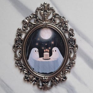 May include: A silver ornate frame with a white oval picture inside. The picture shows two white ghosts sitting at a table with a lit birthday cake. The background is a dark forest with a full moon and stars.
