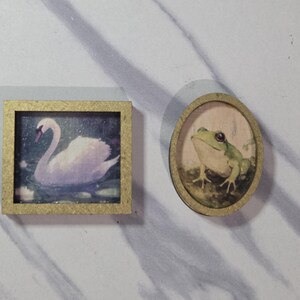 May include: Two miniature framed paintings. One square frame features a swan on water, the other is an oval frame with a frog. Both frames have a gold-colored trim.