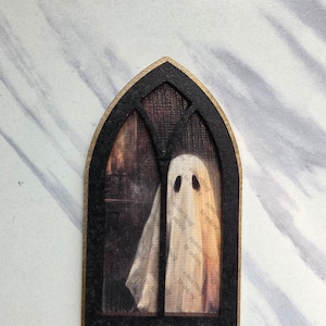 May include: A decorative wall ornament shaped like a pointed gothic window frame in black with gold edges. Inside the frame is an illustration of a white ghost figure against a dark background, with the repeated text something to adore visible.