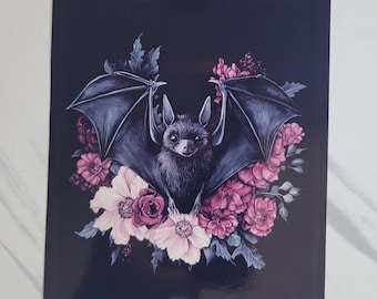 May include: A black bat with pink and white flowers on a dark blue background. The bat is in the center of the image with its wings spread out. The flowers are arranged around the bat in a circle.