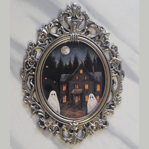 May include: A silver ornate frame with a round glass insert depicting a spooky house with two ghosts in front of it. The house has lit windows and the scene is set at night with a full moon in the sky.