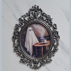 May include: A silver ornate frame with an oval opening, containing a painting of a white ghost sitting at a table with a cup of coffee. The ghost has two black eyes and is wearing a white sheet. The table is made of wood and has a brown surface. The background is a blurry brown and white.