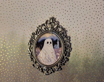 May include: A silver ornate frame with a white ghost in a field of flowers. The ghost has a small mouth and two black eyes. The background is a blue and purple landscape.
