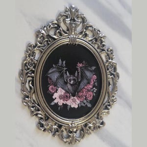 May include: A silver ornate frame with a black background and a detailed illustration of a bat with pink flowers.