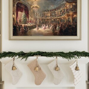 May include: A framed painting of a festive ball scene hangs above a fireplace. Below, four Christmas stockings in neutral tones are suspended from the mantel, each adorned with a small gold bell and a garland of greenery.