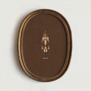 Puede incluir: Un marco de pared ovalado con un acabado dorado ornamentado que contiene un paspartú marrón oscuro. Centrada en el interior hay una pequeña ilustración detallada de un cascanueces con corona, uniforme rojo y dorado, y botas negras.