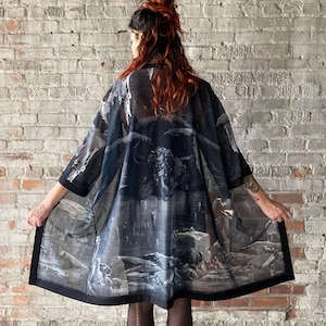May include: A black sheer kimono with a printed design of a dark fantasy scene. The kimono has a black trim and is worn over black leggings and boots.