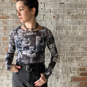 May include: A black mesh long-sleeved shirt with a print of many eyes. The shirt has a ruffled hem and cuffs. The model is wearing black jeans and a silver choker necklace.